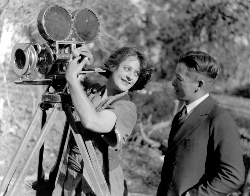 On the left, the film director appears to be inserting film into the camera’s drive mechanism. While doing so, she is looking at a man on the right side of the image.