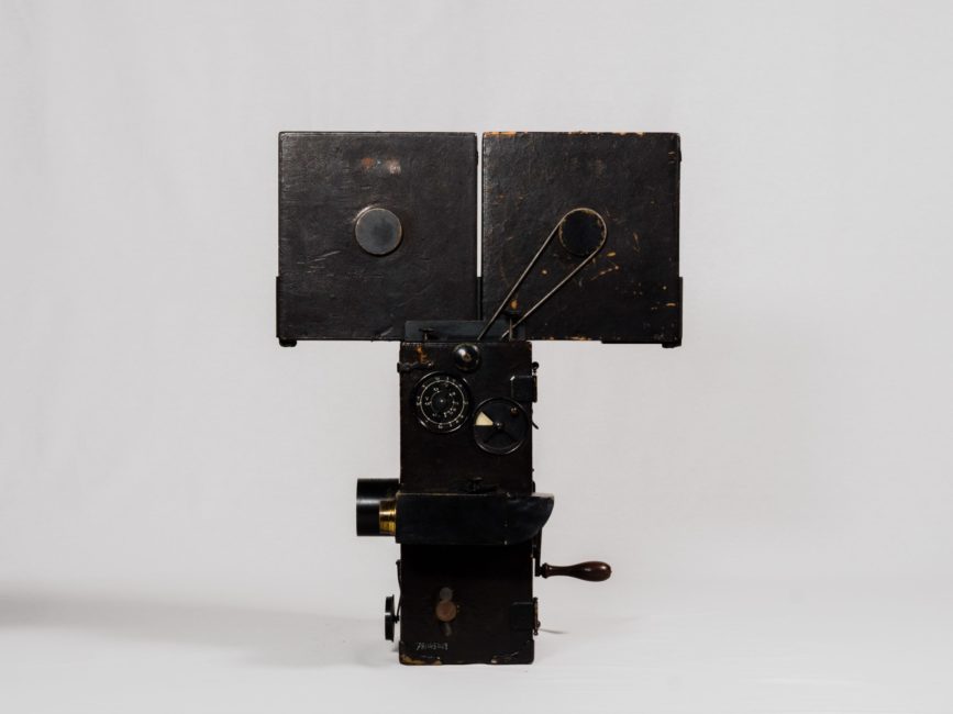 Three-quarter view of the left side of the camera. The device consists of two large black wooden rectangles. One rectangle forms the body of the camera and the other, on top, contains the film. It is operated with a crank located at the rear left of the body.