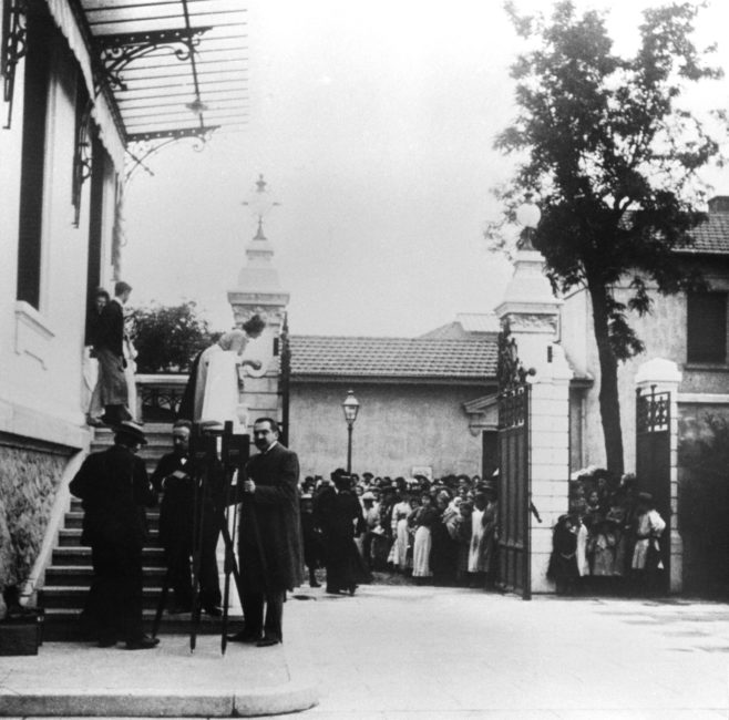 Deux opérateurs non identifiés sont postés à côté de leur Cinématographe, en extérieur devant un grand escalier. À l’arrière-plan, une foule est regroupée devant l’entrée de la propriété.