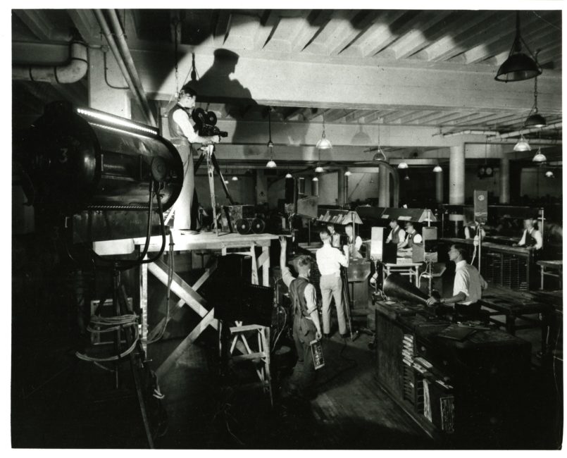Employees work in what appears to be a factory. A camera operator has set up a Bell & Howell on a raised platform. Strong artificial lighting has been added to aid in filming.