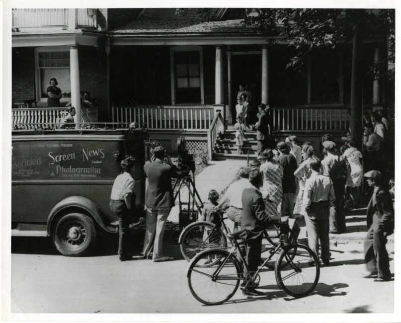 A crowd of onlookers, viewed from behind, is gathered in front of a house. At the left of the image is a truck with “Associated Screen News Photographers” written on it. At the rear of the vehicle, a camera operator and his assistant are filming people on the front porch of the house.