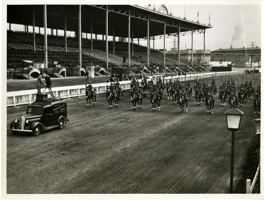 On what appears to be a racetrack, a large number of soldiers are lined up on horseback and moving toward a Bell & Howell camera, which has been set up on the roof of a small van. The camera operator, who is also perched on the roof of the vehicle, is filming the action.