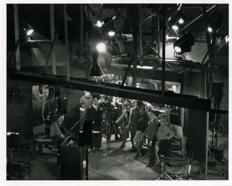 A wide shot of the set. At the top of the photo, lighting is attached to a heavy metal structure. The entire film crew is standing behind the camera, facing one of the actors.