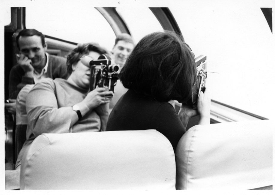 Dans un bus, à droite, Wendy, filme une femme de dos. Joyce filme l’extérieur, à travers une vitre. Deux hommes à l’arrière-plan les regardent en souriant.