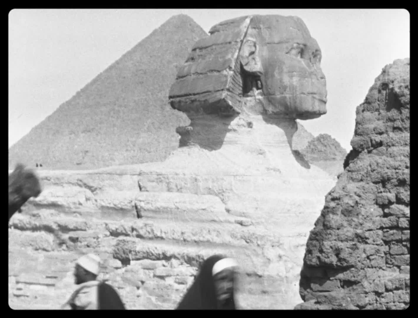 Une caravane de chameliers passe à l’avant-plan. Au centre le Sphinx de Gizeh et à l’arrière-plan apparaît la pyramide de Khéops.