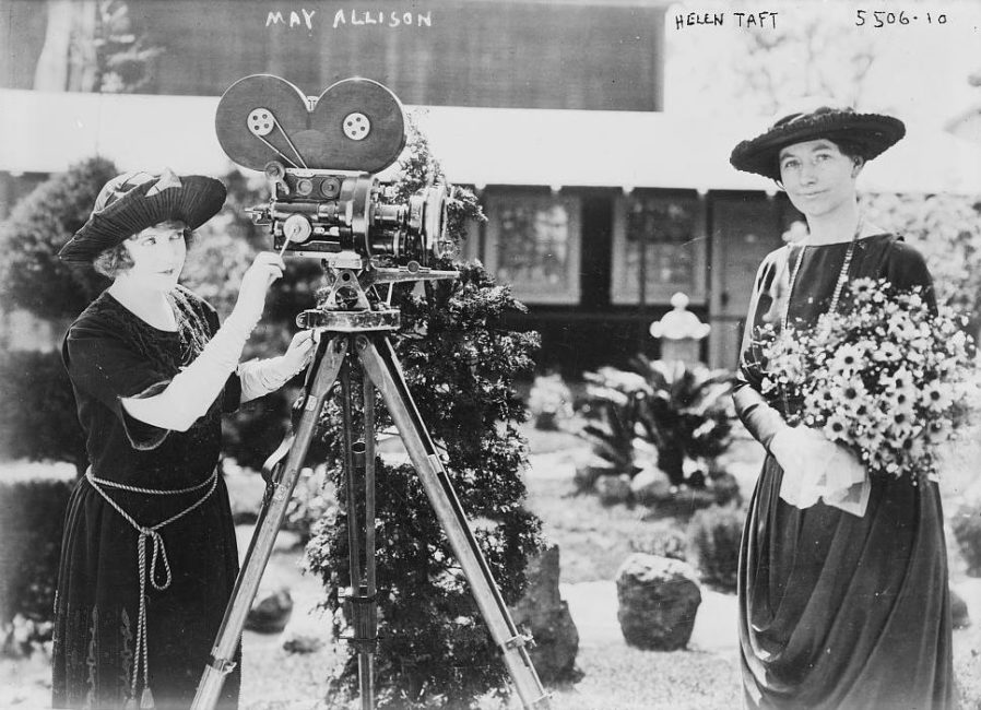 On the left, a woman operates the crank of a Bell & Howell. Another woman is standing in front of the Bell & Howell, looking at the photographer. She is holding a bouquet of flowers. This photograph appears to be staged.