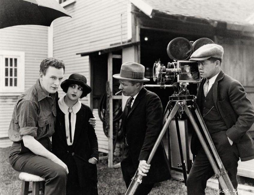 On the left, a man sits on a stool in the shade of an umbrella. He is looking at the photographer. Next to him, a woman is looking at him. They are the actors. On the right, in front of the camera, a man is looking at them while smoking a cigar. Next to him, the camera operator is standing behind the camera with his hand on his hip, observing the scene.