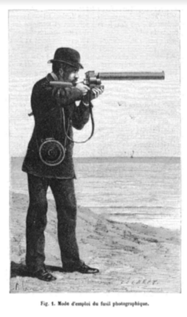 Un homme debout devant la mer vise un élément hors champ avec son fusil. Il a accroché en bandoulière, une boîte conservant les plaques sensibles à l’abri de la lumière.