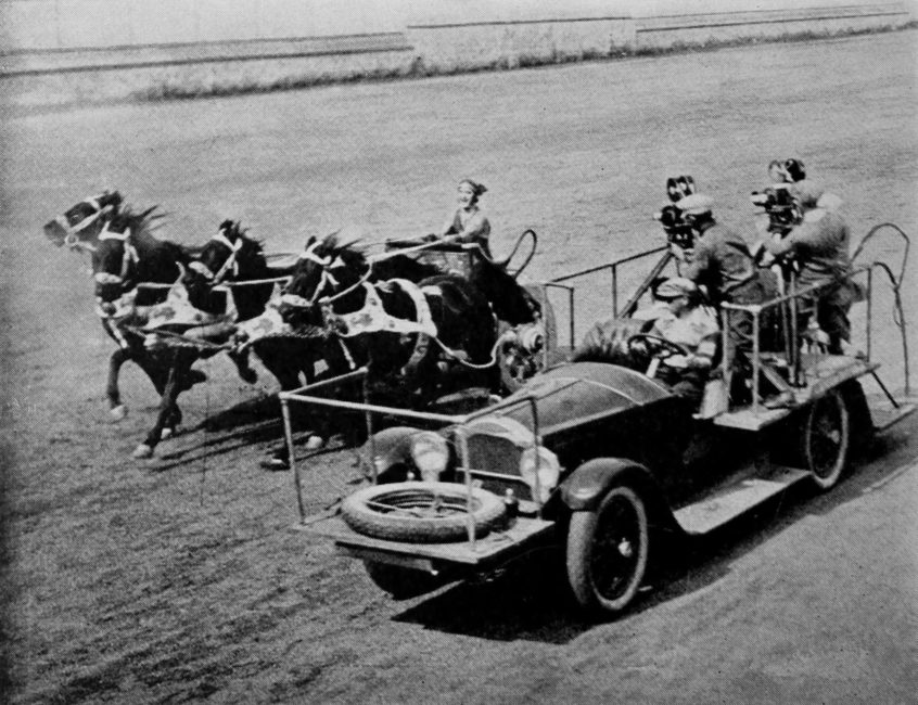 Wide view. In the background, left side, a chariot is being pulled by four galloping horses. On the right, on a car fitted out with a platform, are two cameramen, side by side. They are filming the race.