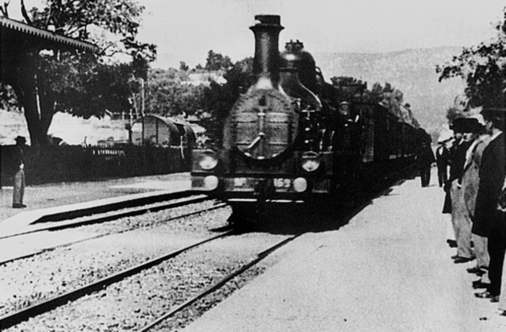 Le train arrive en gare. Les voyageurs à droite du cadre attendent sur le quai.