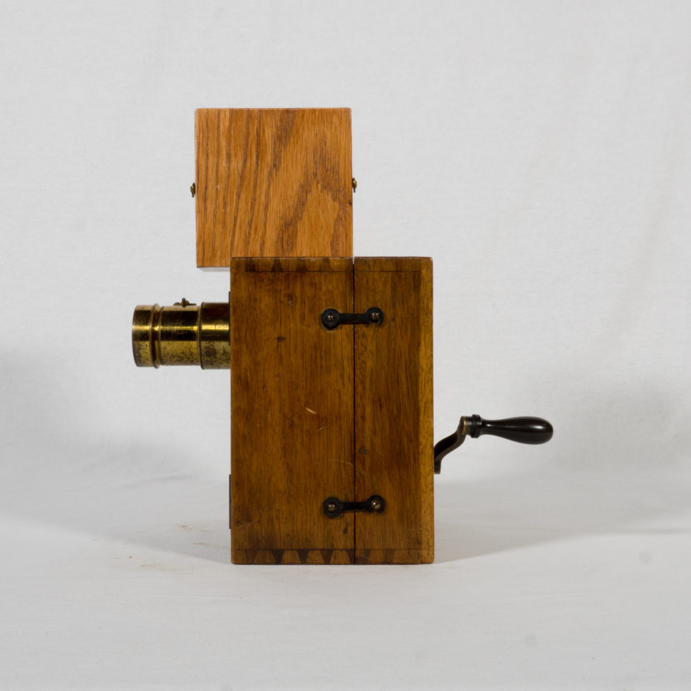 On aperçoit le corps carré de la caméra en bois surmonté d’une plus petite boîte qui contient la pellicule vierge. L’objectif est visible à l’avant et la manivelle à l’arrière. Deux petits crochets sont positionnés sur le côté et permettent de maintenir la caméra fermée.