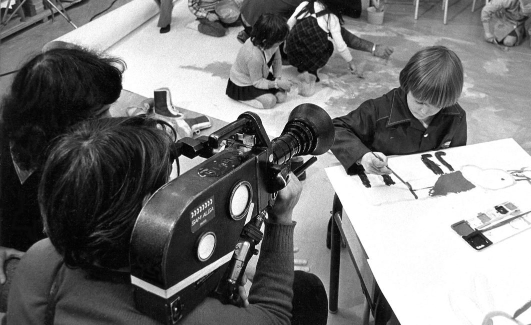Un cameraman filme avec la caméra Éclair, semblable à la Aaton, des enfants en train de colorier.
