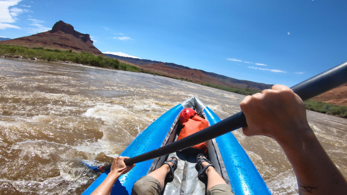 Le bout d’un kayak ainsi que les bras et les jambes de la kayakiste sont visibles. Elle est au milieu d’une rivière à la couleur marron. Au loin, on voit une montagne.